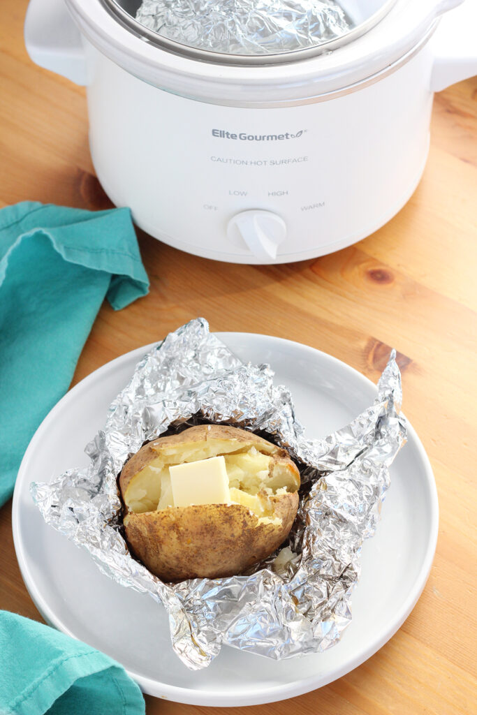 image showing a wooden table top with a small white crock pot off in the background with a teal napkin. There is  a small round white plate with a baked potato that is wrapped in foil partially and has a tab of butter on top