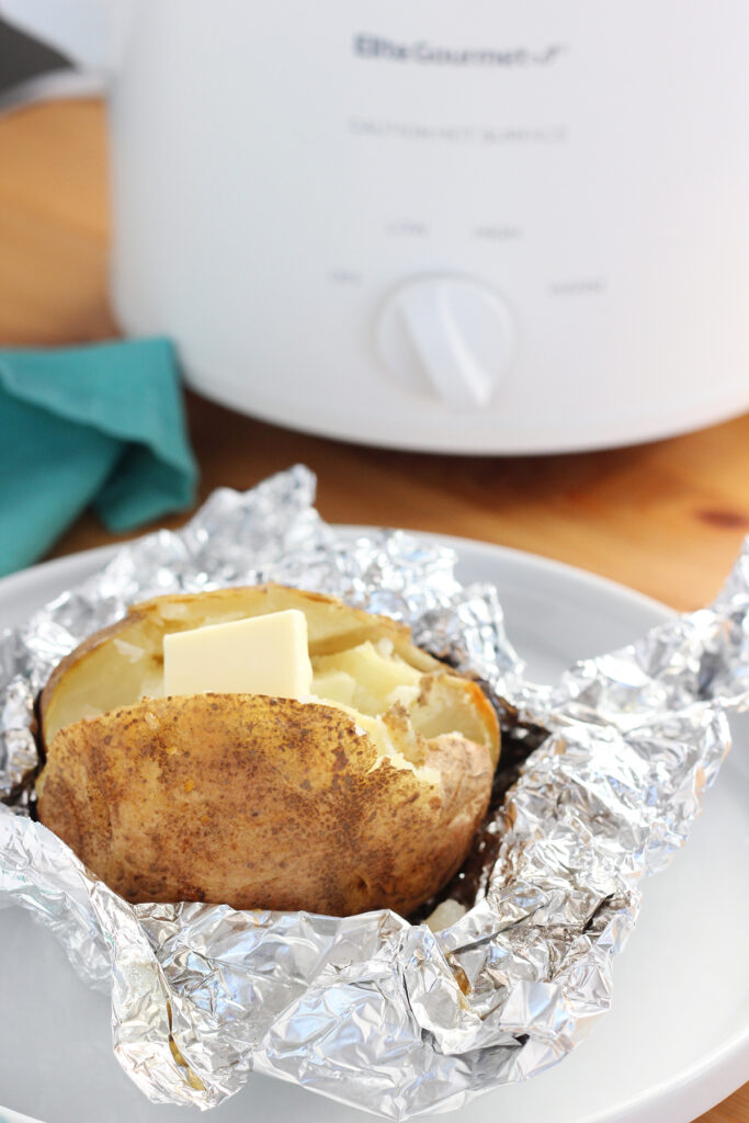 image showing a wooden table top with a small white crock pot off in the background with a teal napkin. There is  a small round white plate with a baked potato that is wrapped in foil partially and has a tab of butter on top