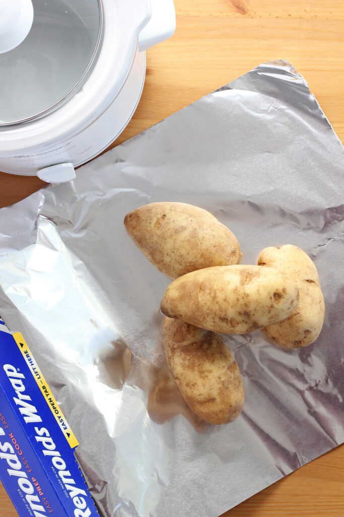 top down image showing a sheet of tin foil being pulled from a reynolds wrap container. On top of the foil are four potatoes. There is a white slow cooker off to the side