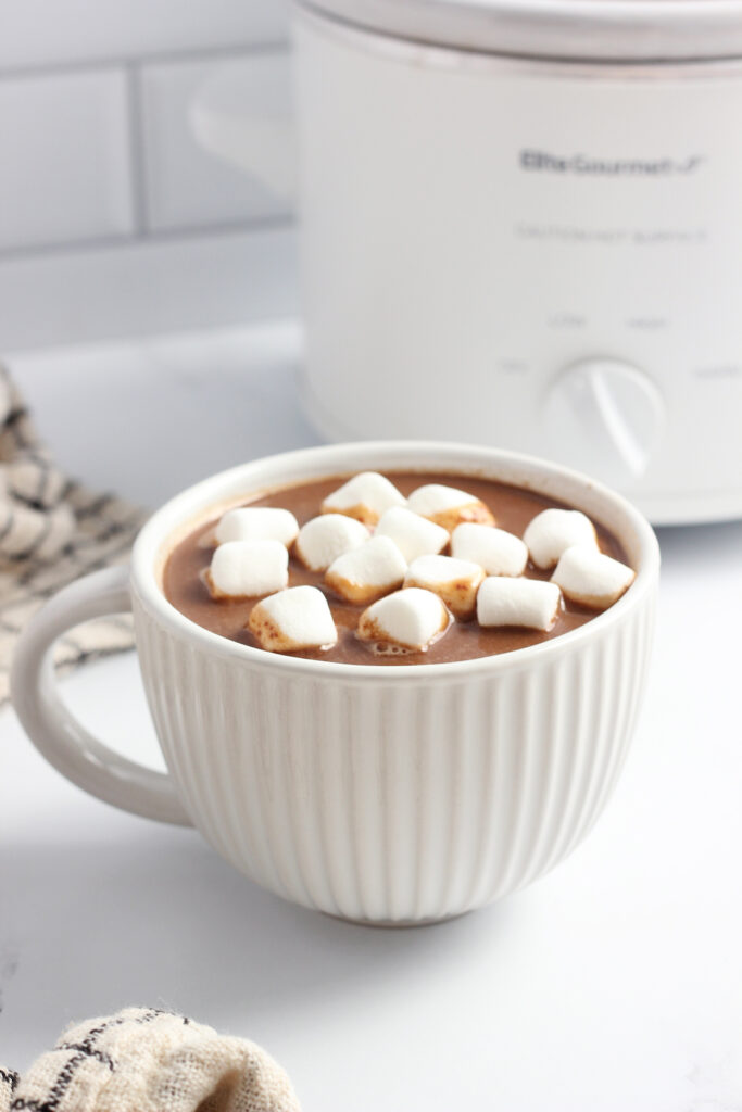 close up image of a white mug that is lined with ridges and filled with cocoa with marshmallows on top. The cup is sitting on a white table top with a white slow cooker off in the distance