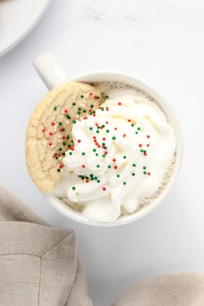 top down image showing a white mug that is filled with coffee, whipped cream, half a sugar cookie, and red and green holiday sprinkles. The mug is sitting on a white table top with a white round plate with more cookies and a tan napkin off to the side