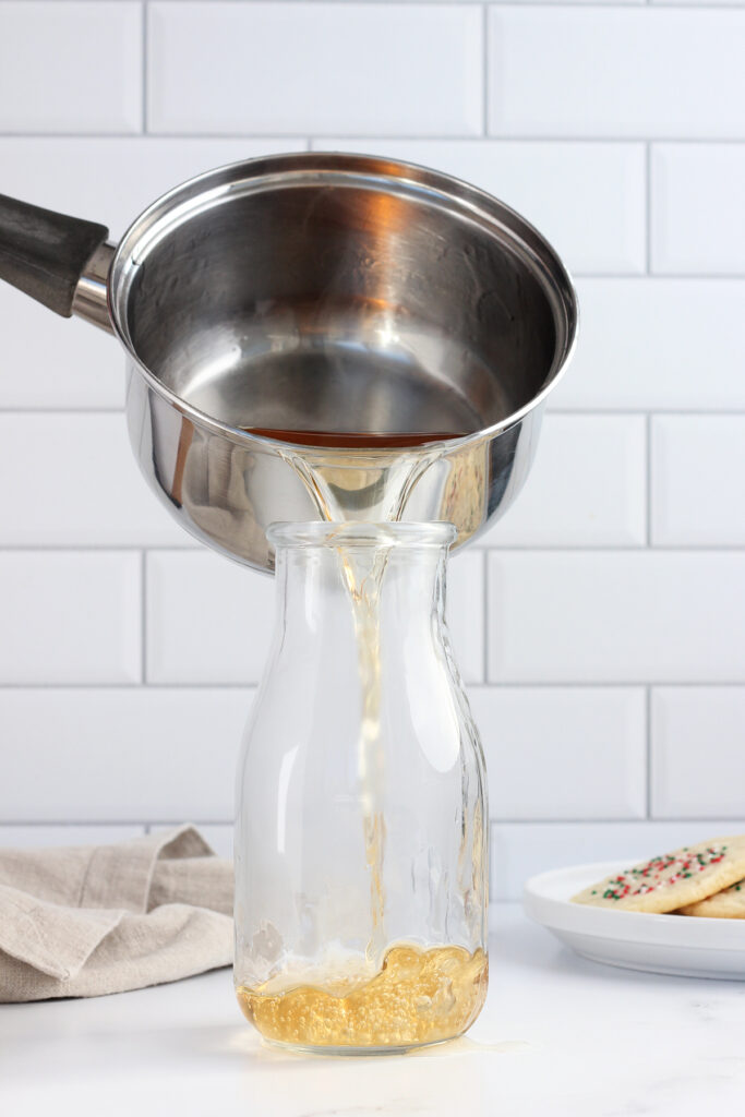 syrup being poured from a metal pan into a tall glass jar. The jar is sitting on a white table top with a small round plate of cookies and a tan napkin off to the background