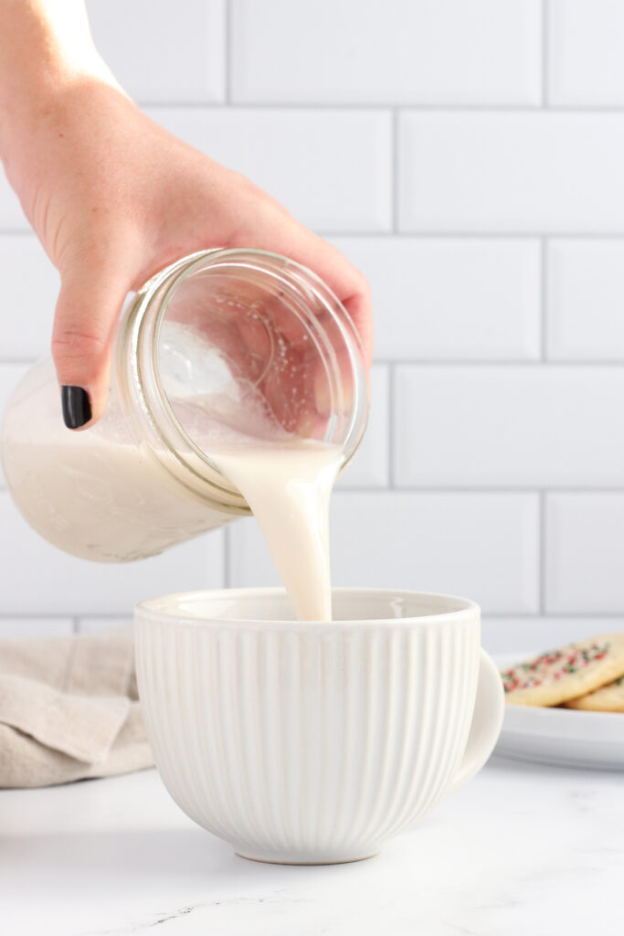 white ridges mug sitting on a white countertop with a plate of sugar cookies off to the side. A hand is holding a mason jar and pouring frothed milk into the glass