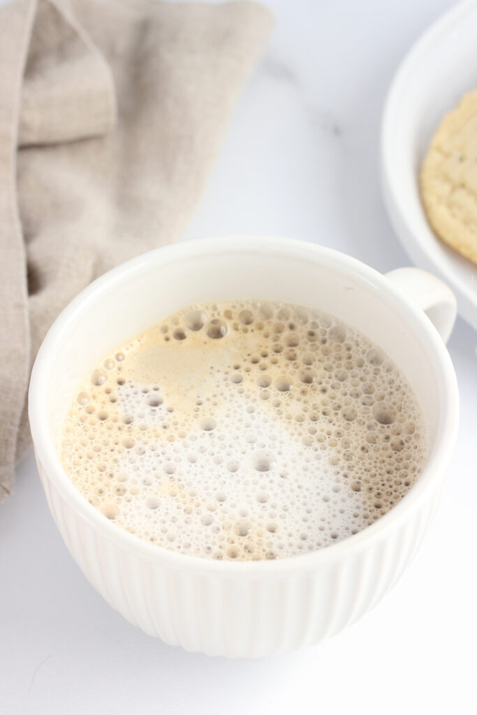 close up image to a white mug that is sitting on a white table top with a teal napkin off to the side. Inside the mug is a latte 