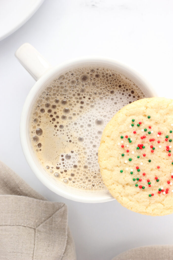 top down image that is close up to a white mug that is sitting on a white table top with a teal napkin off to the side. Inside the mug is a latte with a sugar cookie balancing on the edge of the glass