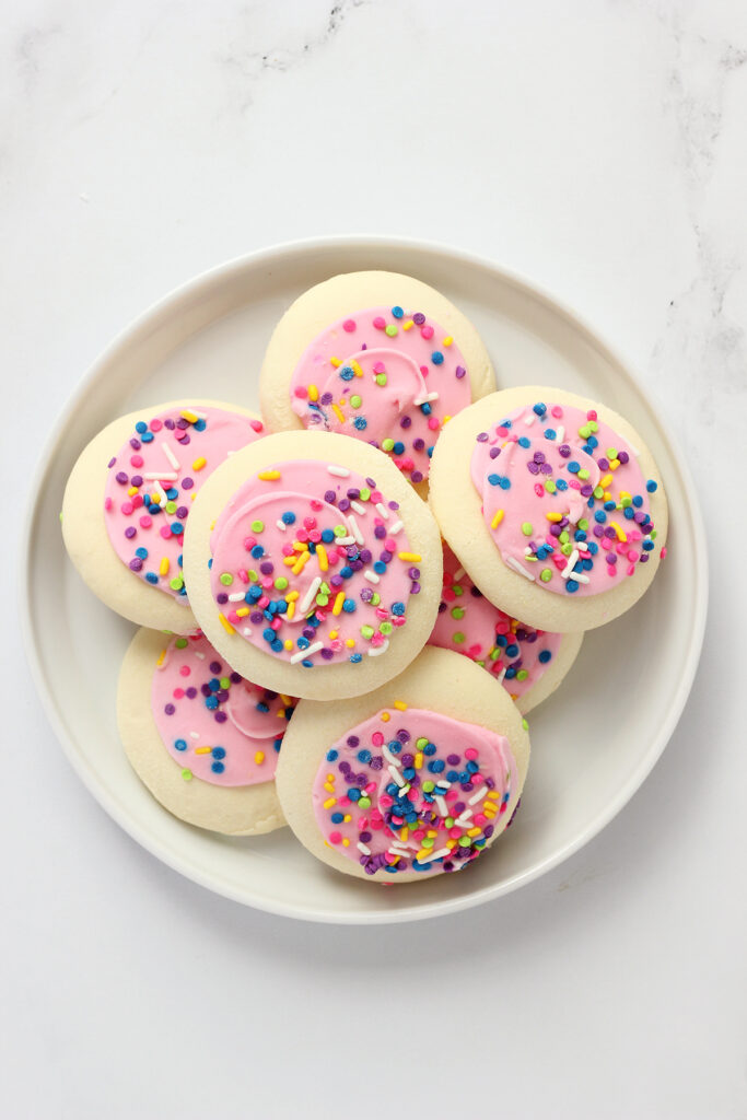 top down image showing a small white plate with a lifted edge sitting on a white table top. The plate is stacked with store bought sugar cookies that are frosted in a pink frosting and rainbow sprinkles