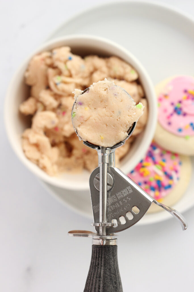 top down image focusing on a cookie scoop that is filled with a sugar cookie dough. The scoop is being held over the top of a small whiten bowl with more dough and two sugar cookies off to the side. 