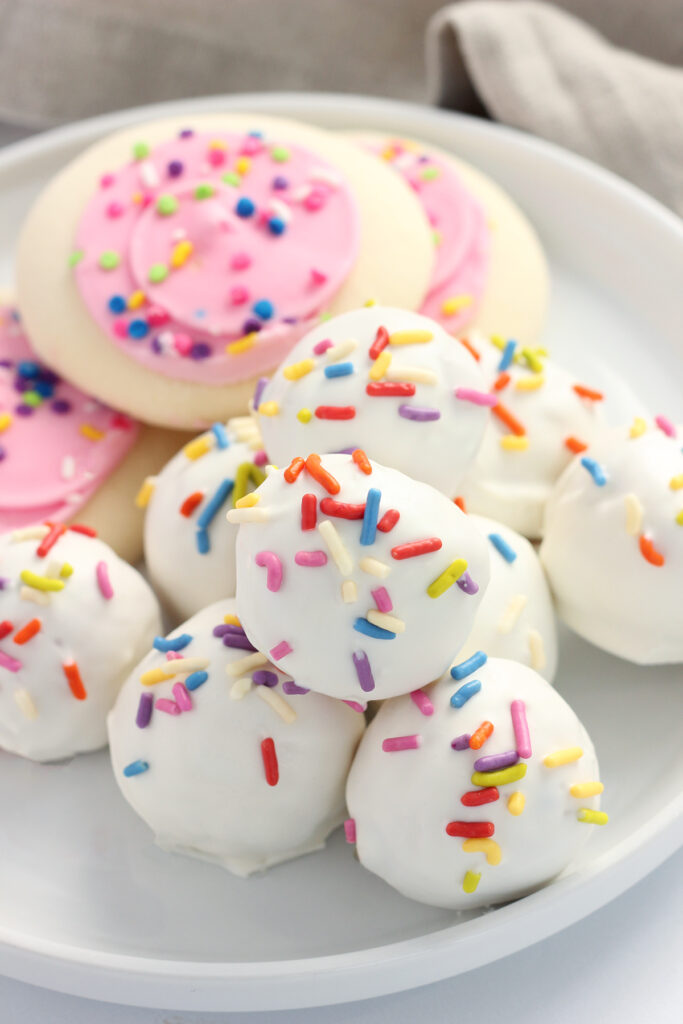 close up showing a stack of white truffles with rainbow sprinkles sitting on a white plate with a lifted edge. Pink frosted sugar cookies are off in the background