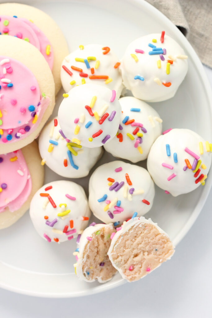 top down image showing a plate of white truffles with rainbow colored sprinkles sitting on a white round plate. There are three sugar cookies off to the side stacked and one truffle cut in half showing the inside