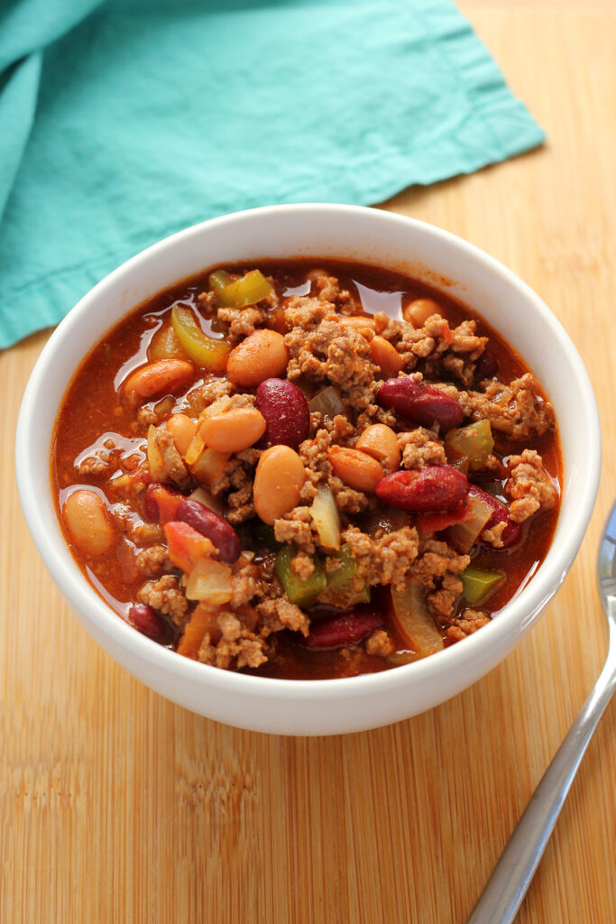 image showing a white bowl filled with chili. The bowl is sitting on a wooden table top with a teal napkin off to the side