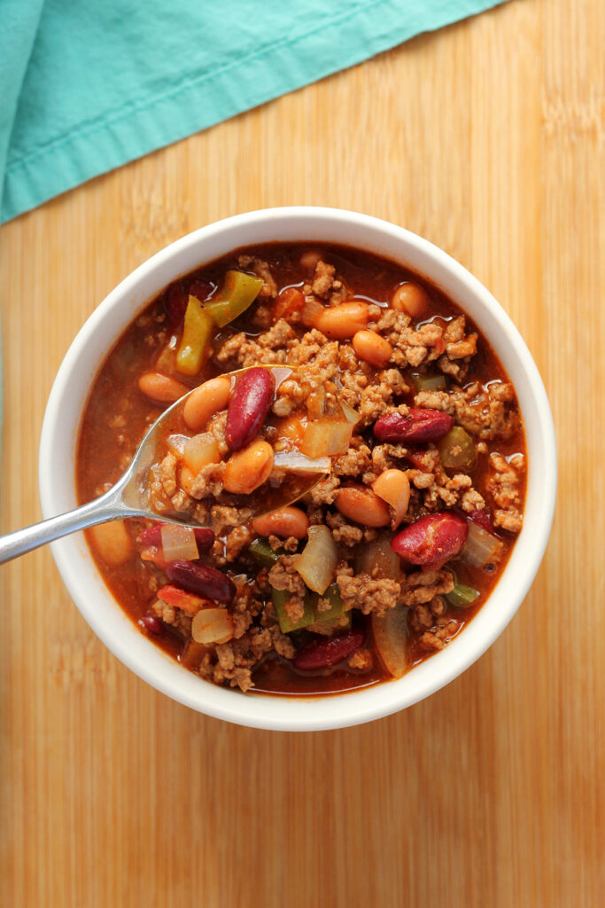 top down image showing a white bowl filled with chili. A bite is on a spoon hovering over the top of the bowl. The bowl is sitting on a wooden table top with a teal napkin off to the side