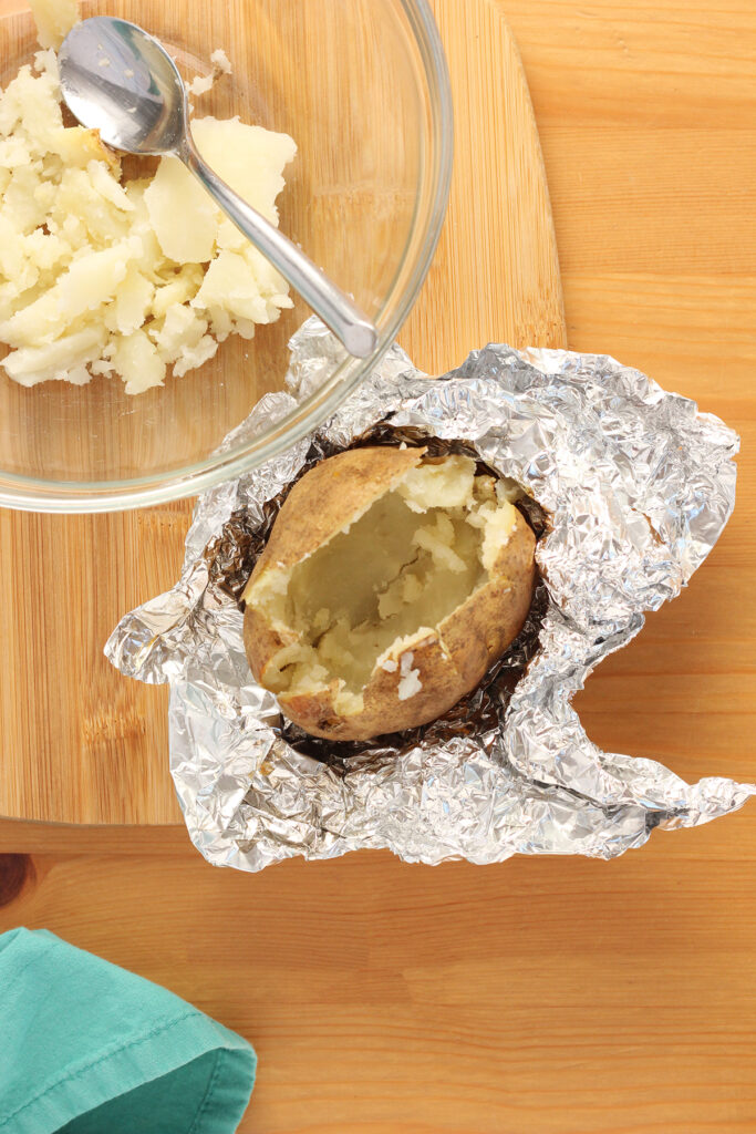 top down shot showing a baked potato sitting in a foil wrapping with the center scooped out into a glass mixing bowl
