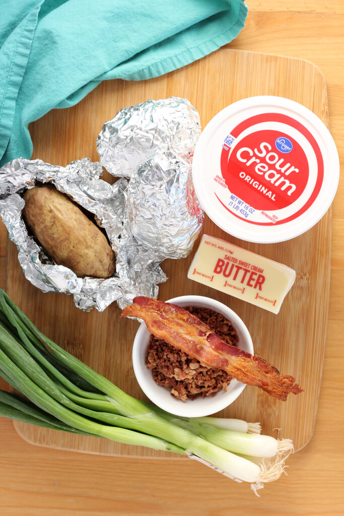 top down image showing a wooden cutting board with toppings for a baked potato. This includes cooked potatoes wrapped in foil (one that is slightly unwrapped), a tub of kroger sour cream, a stick of salted butter, a small white bowl filled with bacon crumbles and a slice of bacon hovering on the edge of the bowl, and a bundle of fresh green onions