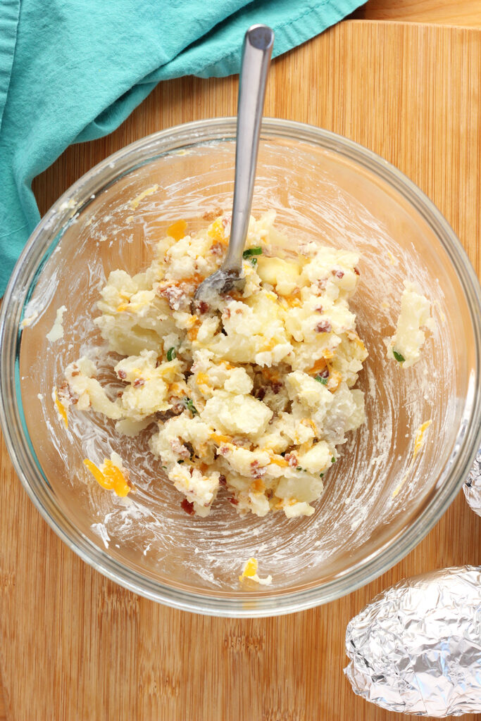 top down shot showing a glass mixing bowl filled with soft potato, pat of butter, scoop of sour cream, and a sprinkle of bacon, cheese, and green onion slices mixed together