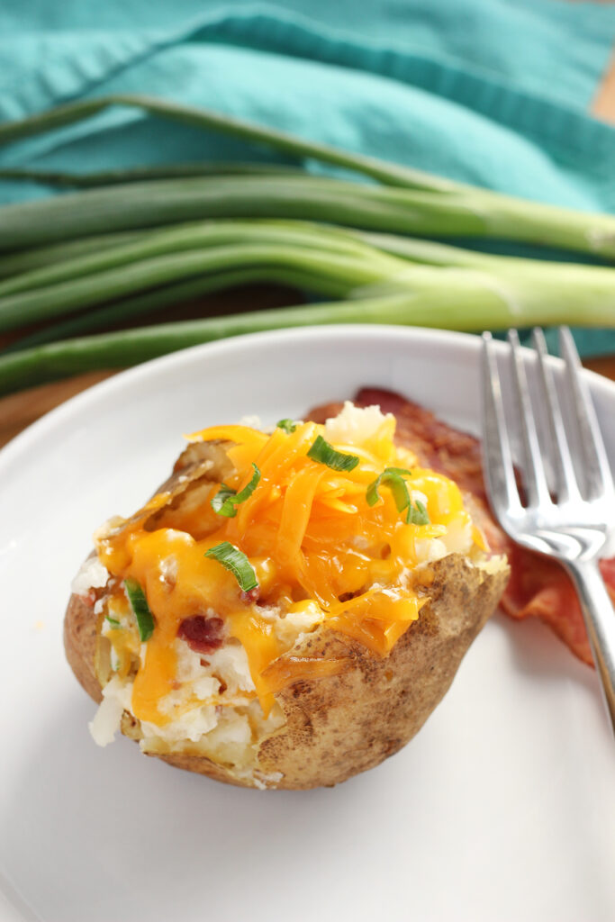 close up image showing a potato that has been sliced in half and filled with cheese, bacon, and green onion. The potato is sitting on a white plate that is round with a slightly lifted edge with a slice of bacon and fork off to the side