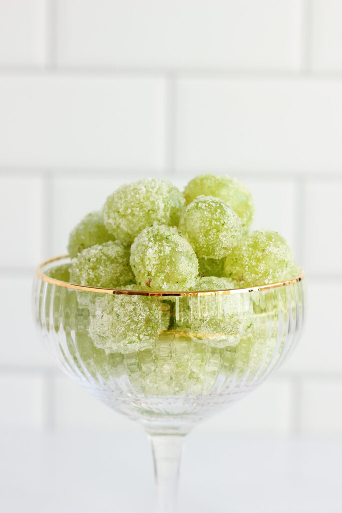 close up image showing a champagne glass that is lined with gold. The glass is filled with sugar coated green garpes