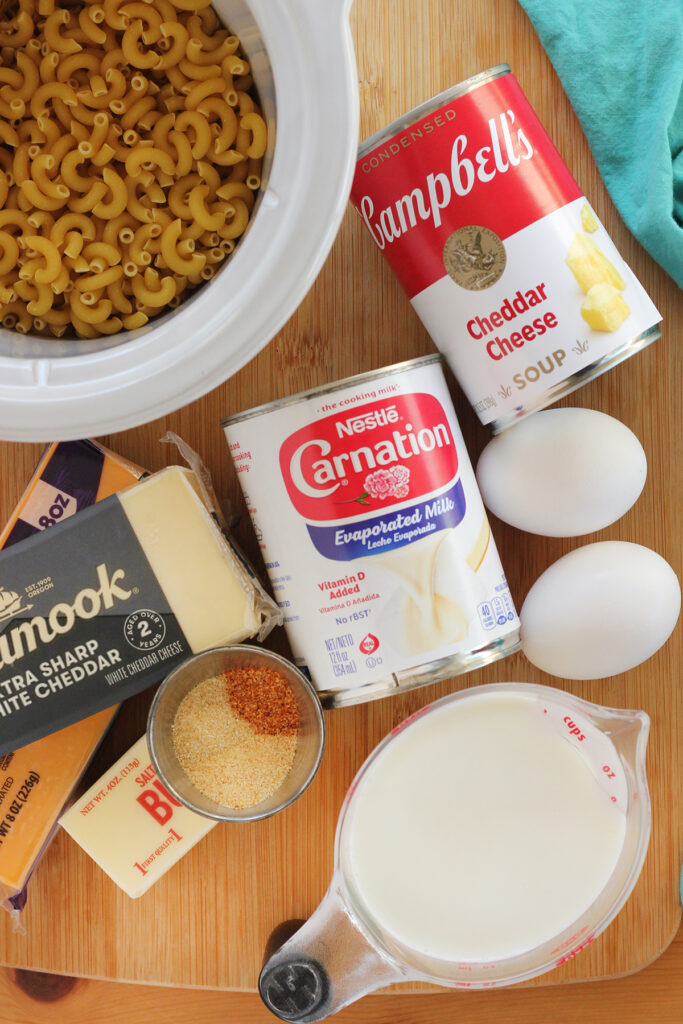 top down image showing a wooden table top that is filled with incredients for macaroni and cheese. There is a small slow cooker container that is filled with uncooked macaroni noodles, a can of Campbell's cheddar cheese soup, a can of Nestle carnation evaporated milk, two blocks of  cheese, a stick of butter, a cup of milk, and a small container of spices