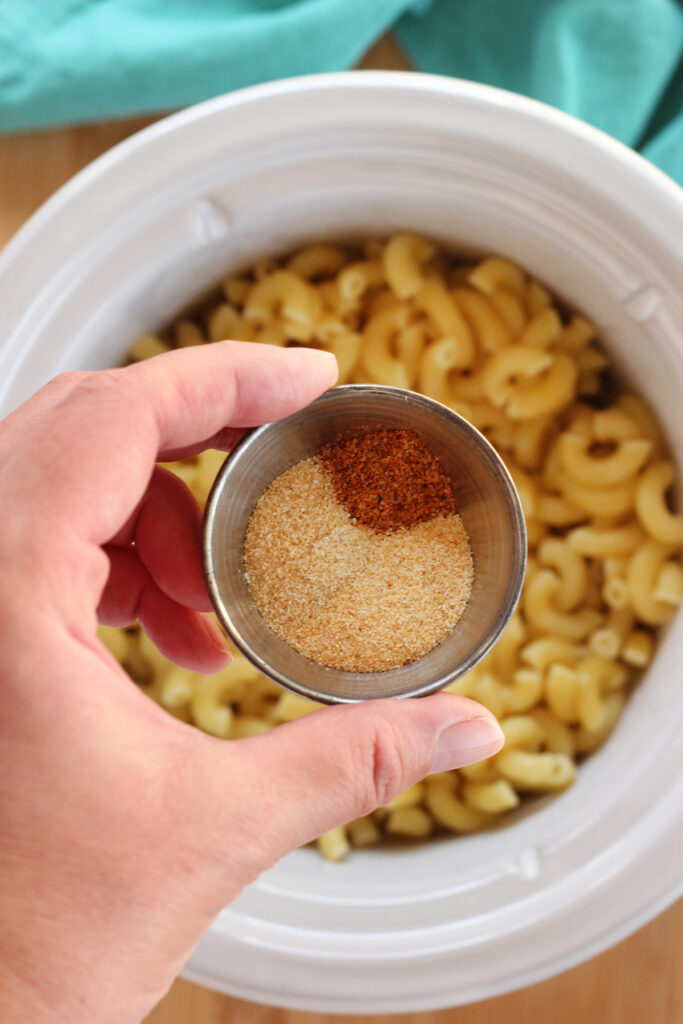 top down image showing a small tin filled with spices being held over the top of a small round slow cooker that is filled with noodles. 
