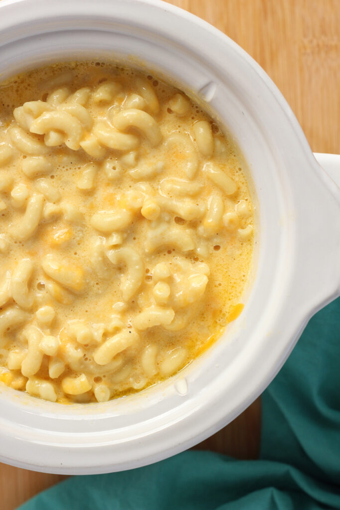 close up top down image showing a slow cooker filled with macaroni and a runny cheese sauce. The slow cooker is sitting on a wooden cutting board with a teal napkin and a plate of orange and white cheese off to the side. 