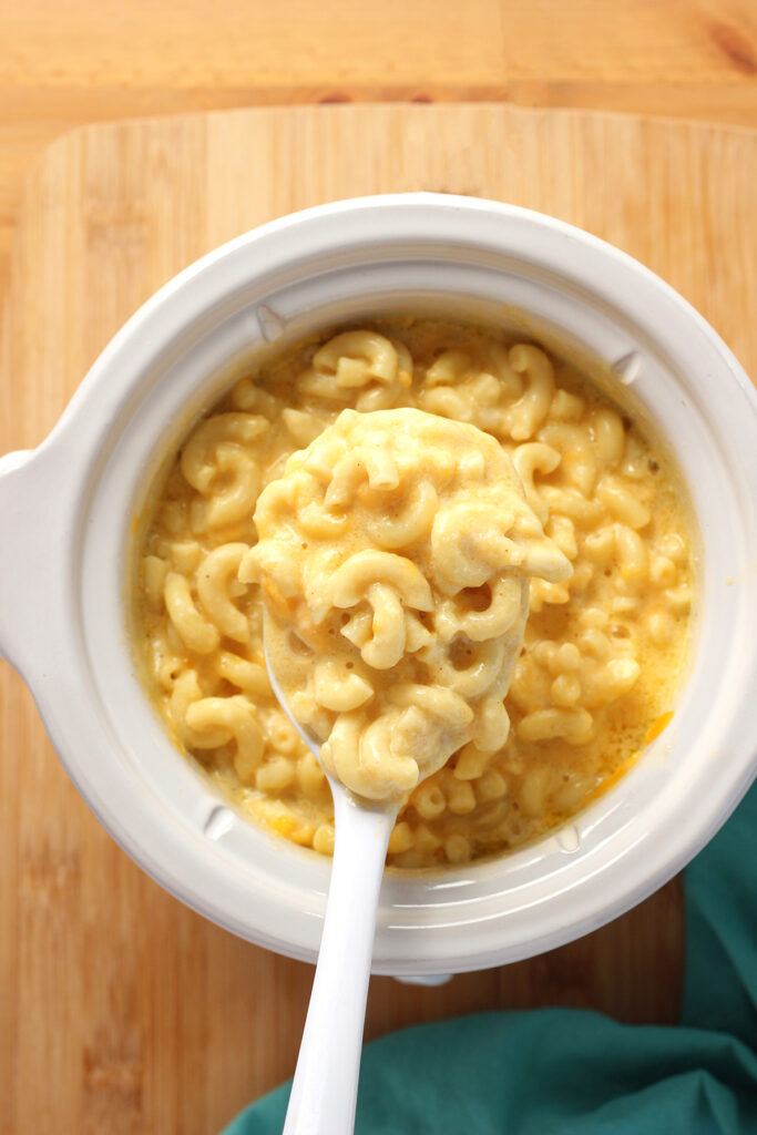 top down image showing a small slow cooker filled with macaroni and cheese. A white spoon scooped a helping out and is hovering over the top of the container. The slow cooker is sitting on a wooden table top with a teal napkin sitting off to the side