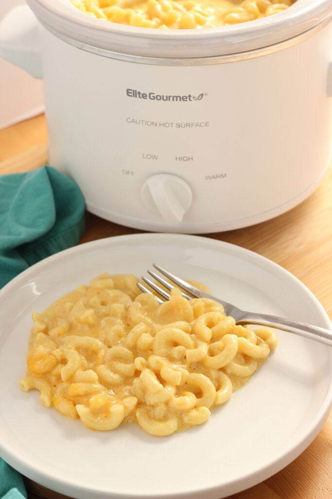 image close up to a white round plate that has a helping of macaroni and cheese with a fork. The plate is sitting on a wooden table with a teal napkin and a small slow cooker off to the side in the background