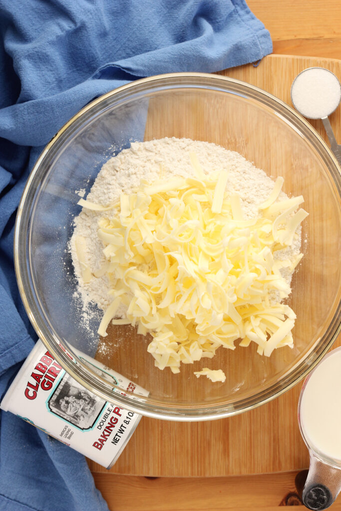 top down image of a large glass mixing bowl filled with flour on a wooden table with shredded butter