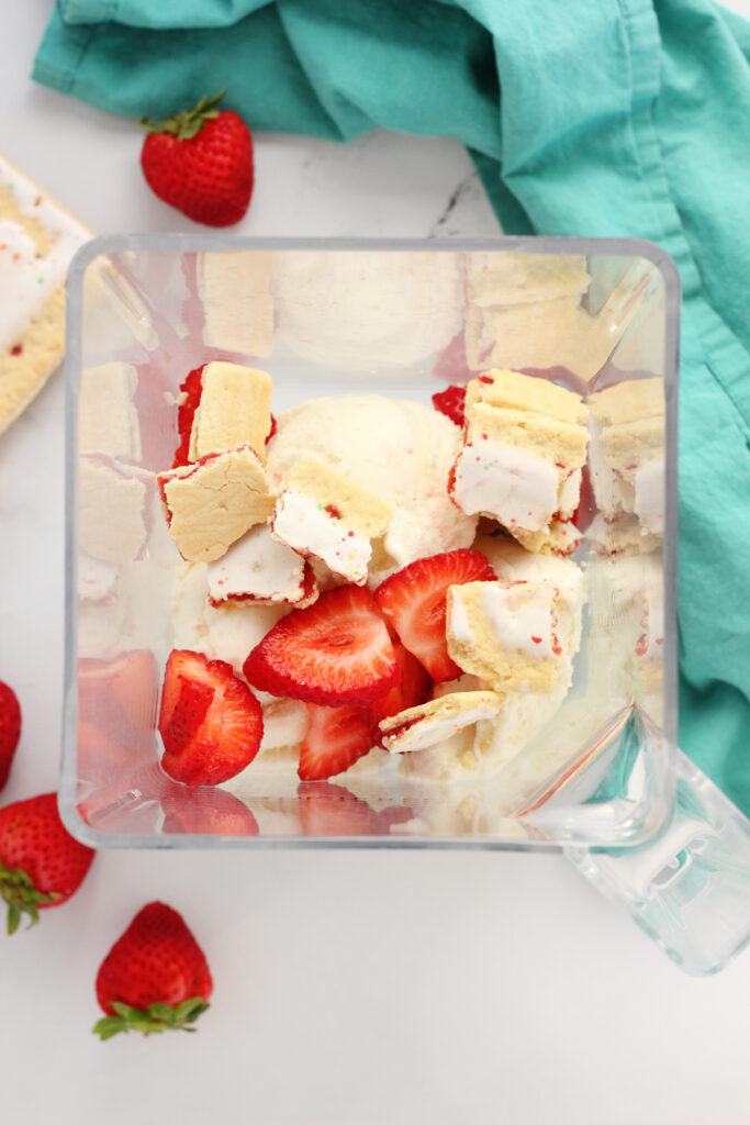 top down image showing blender with the lid off. Inside are a few scoops of vanilla ice cream,  strawberries, and pop tart pieces. Around the container is a pop tart, four strawberries, and a teal napkin