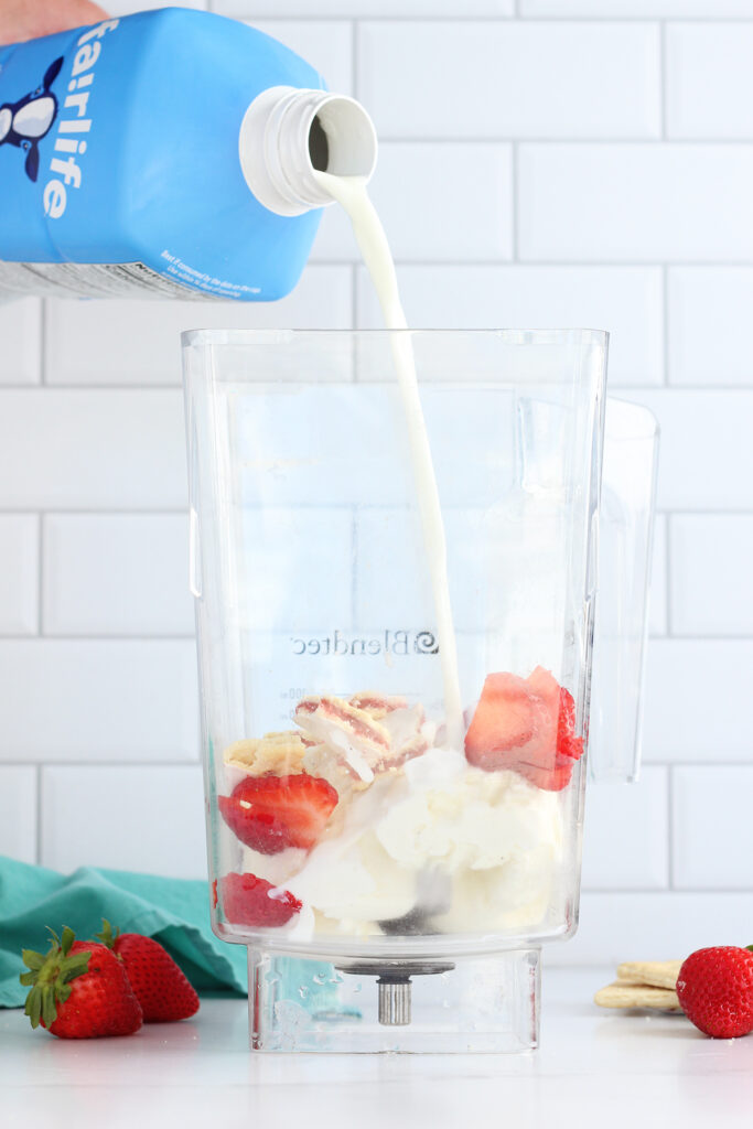 hand holding a blue container of fairlife milk that is being poured into a blender that has vanilla ice cream, strawberries and pop tart pieces. The blender is sitting on a white table top with fresh berries and a teal napkin