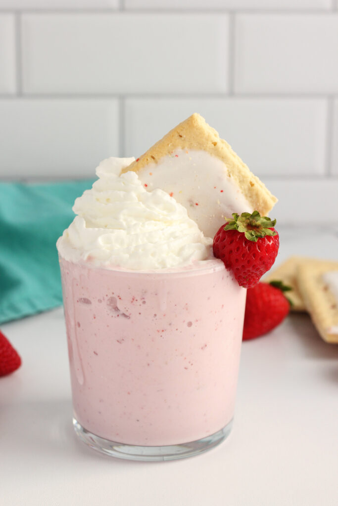 close up image showing a short glass filled with a pink milkshake. The milkshake is topped with whipped cream, a fresh strawberry hanging off the side of the glass, and piece of pop tart in the whipped topping. The glass is on a white table top with a teal napkin off in the distance