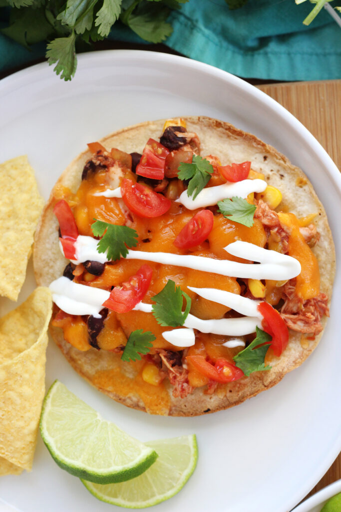 close up image showing a corn tortilla laying flat on a white round plate. On top of the tortilla is shredded chicken, melted cheese, sour cream, tomatoes, and cilantro