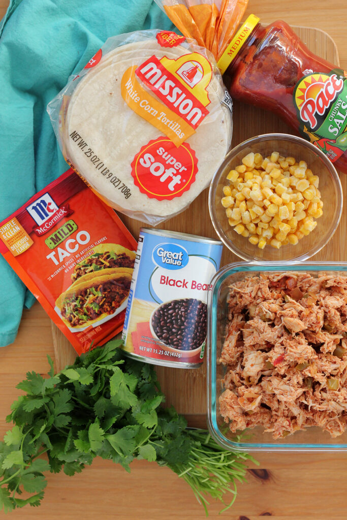 top down image showing ingredients on a wooden table top with a teal napkin off to the side. Ingredients include a square clear dish with shredded chicekn, a bushell of fresh cilantro, a can of great value black beans, a packet of mild taco seasoning, a small glass bowl of frozen corn, a stack of mission white corn tortillas, and a container of paces chunky salsa