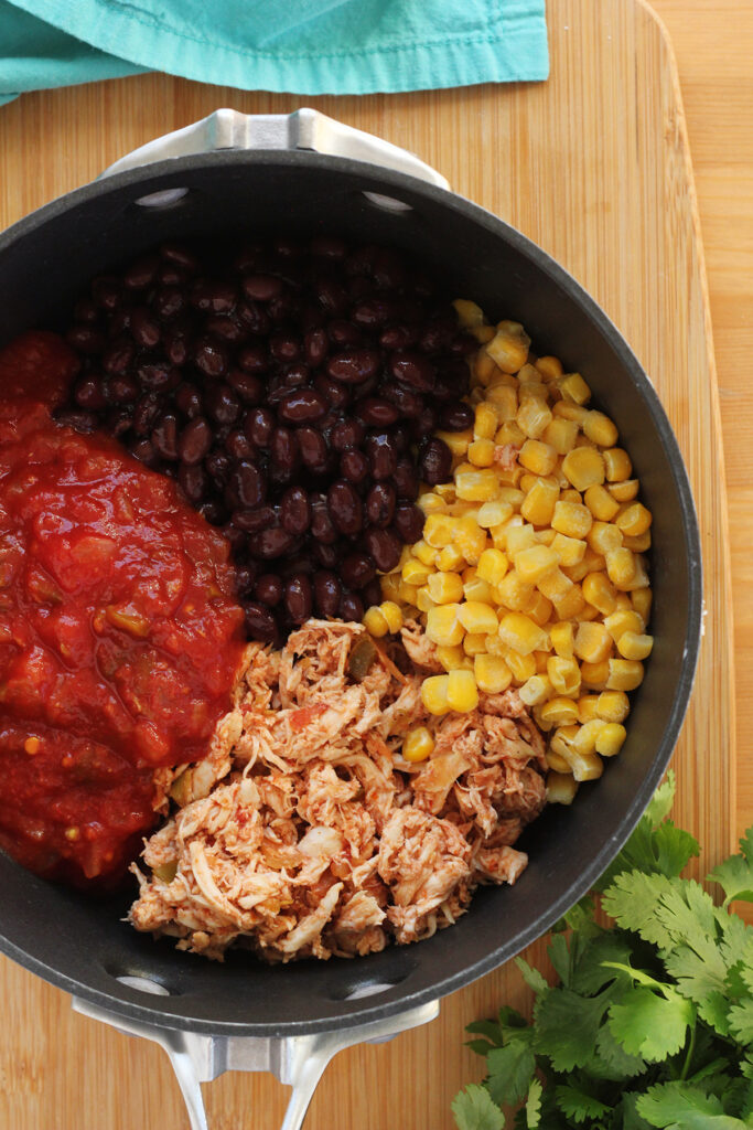 top down image showing a close up shot of a black pan filled with shredded chicken, corn, black beans, and salsa on a wooden table top