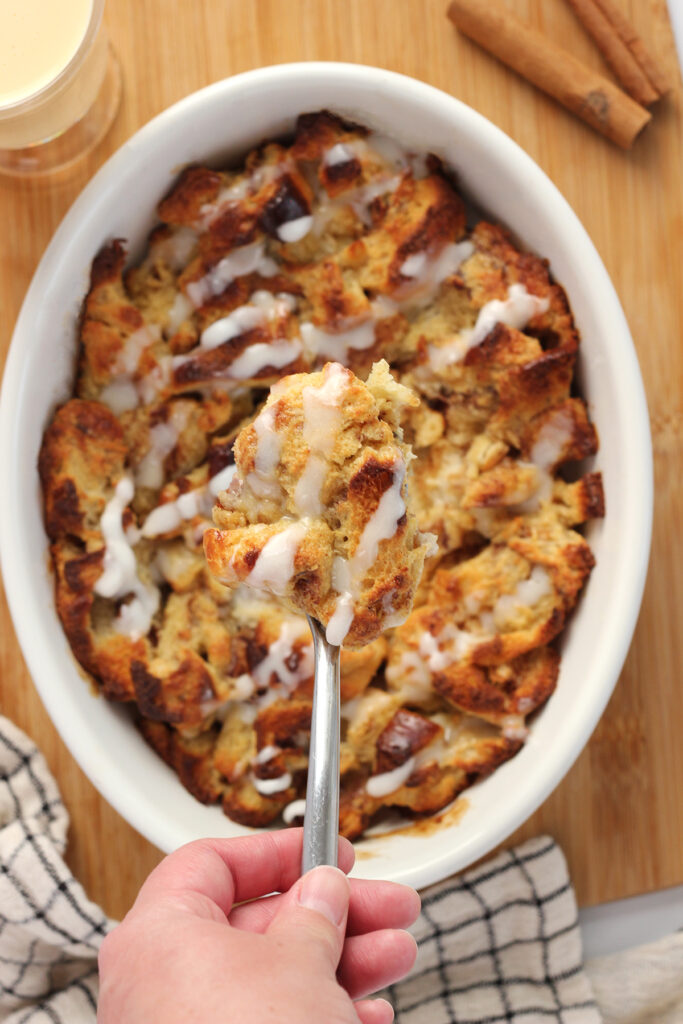 top down shot showing a white oval baking dish filled with cooked bread pudding. This dish is sitting on a wooden cutting board with a white and black checkered napkin, two cinnamon sticks, and a cup of eggnog off to the side. A spoon is holding a bite over the top of the dish