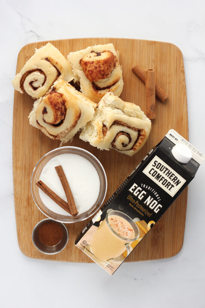 top down image showing a wooden cutting board sitting on a white table top. On top of the board are four baked cinnamon rolls, a glass bowl filled with sugar topped with two cinnamon sticks, a small metal conatiner with brown spices, and a carton of southern comfort eggnog