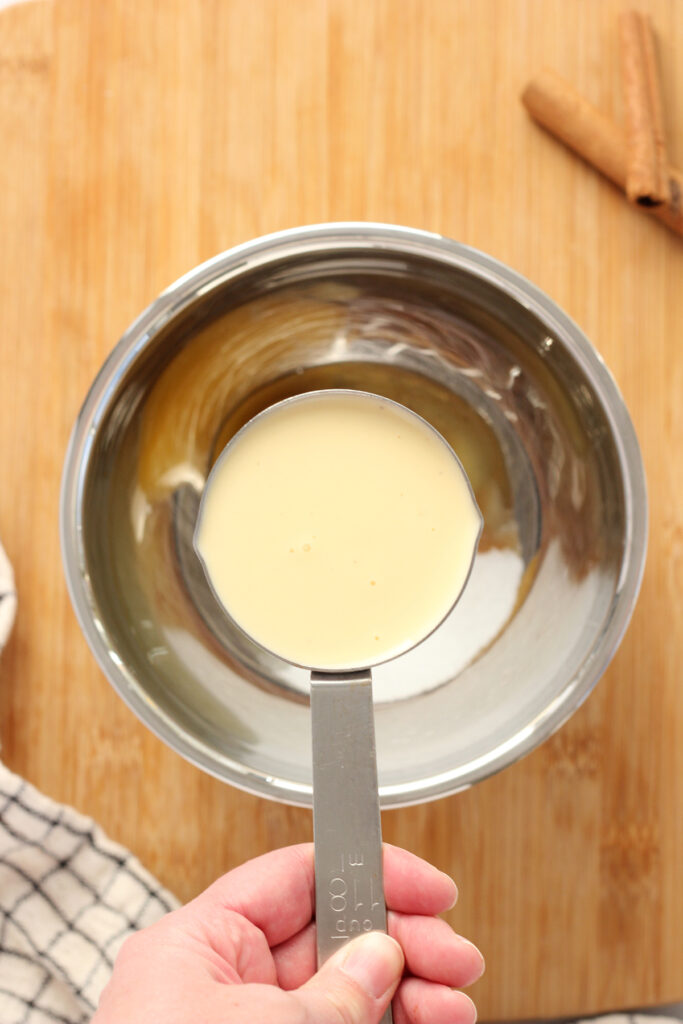 top down image showing a metal mixing bowl with a hand holding a metal measuring cup filled with eggnog over the top