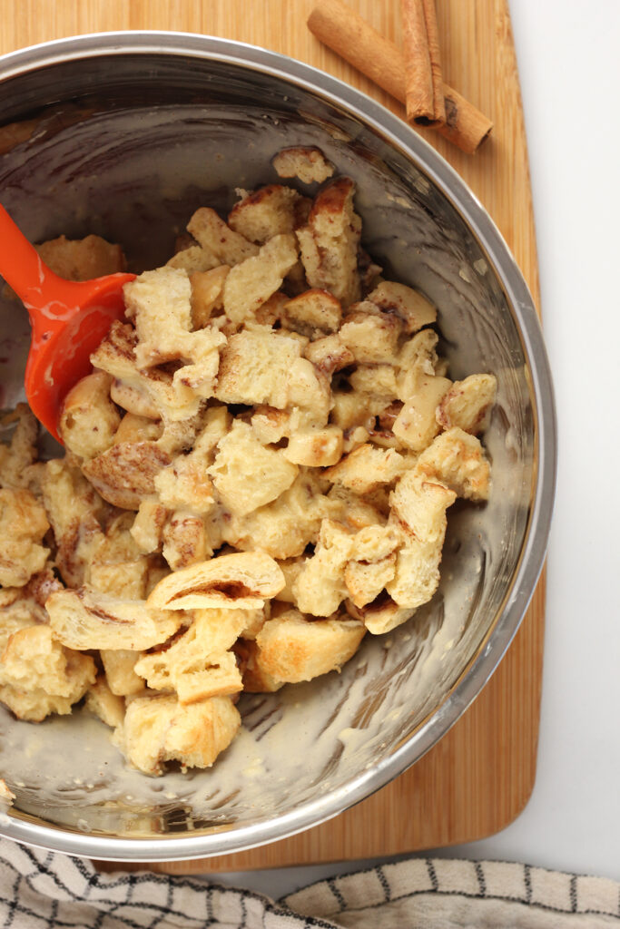 close up image showing a metal mixing bowl filled with cut up bread pieces that have been mixed with an eggnog mixture. 