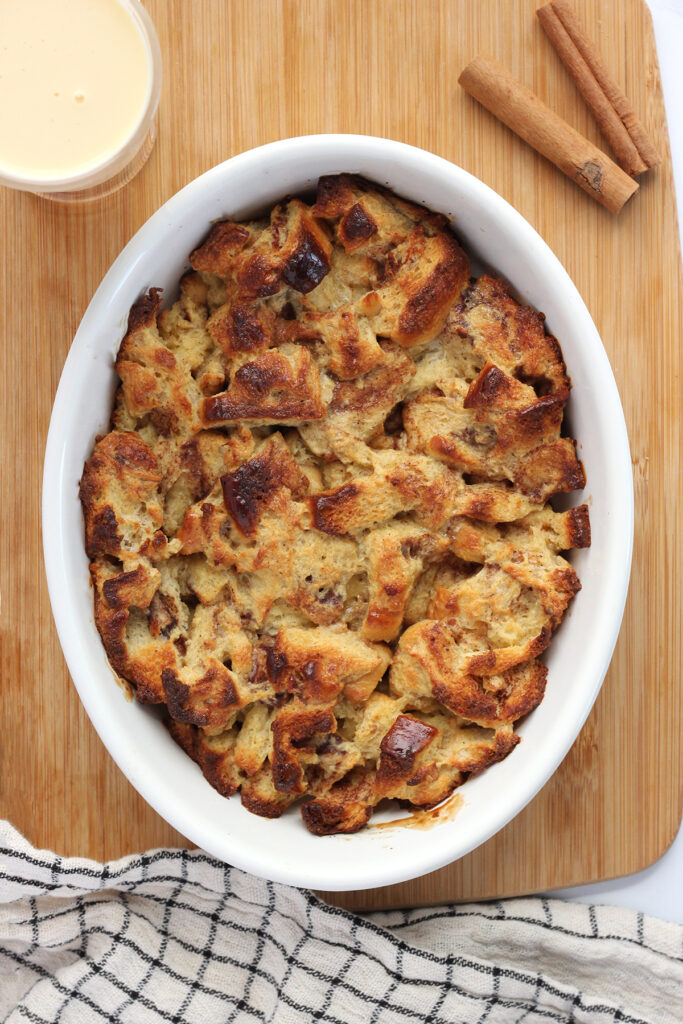 top down shot showing a white oval baking dish filled with cooked bread pudding. This dish is sitting on a wooden cutting board with a white and black checkered napkin, two cinnamon sticks, and a cup of eggnog off to the side