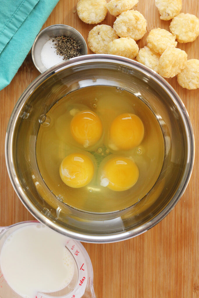 top down image showing a metal bowl filled with four eggs on top of a wooden cutting board which has a measuring cup of milk, frozen tater tots and a bowl of seasonings with a teal napkin
