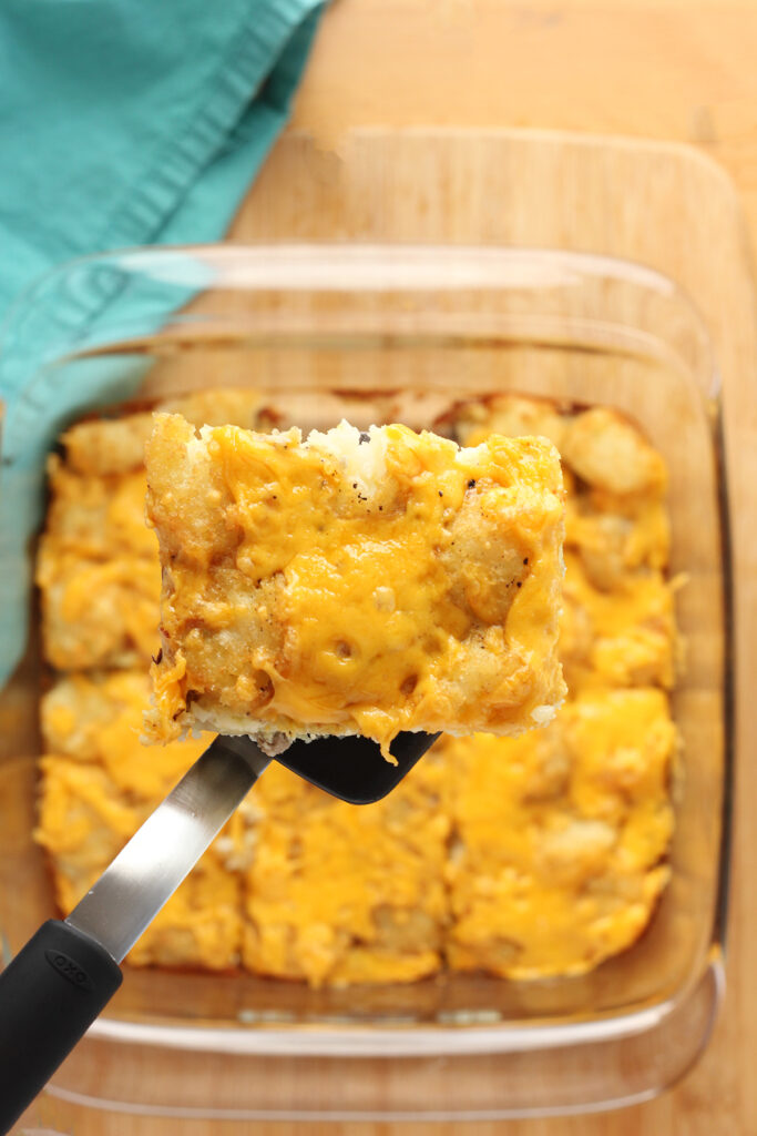 top down image showing a casserole dish filled with eggs, tater tots and cheese. Hovering over the top is a spatula holding a serving of the egg