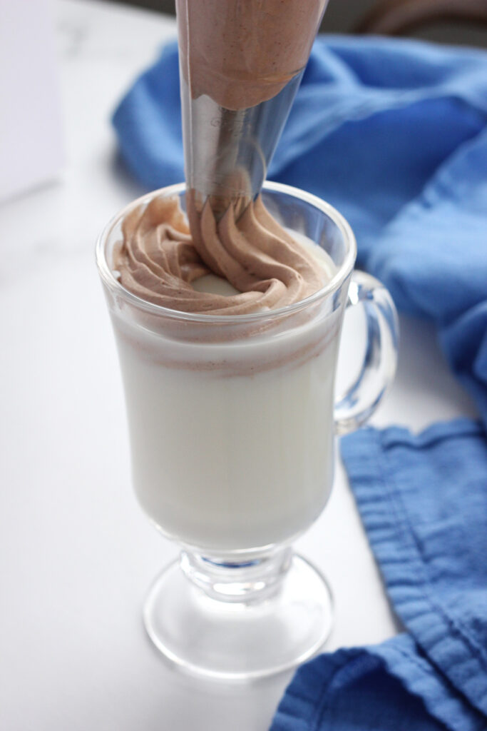 close up image showing a glass mug filled with chocolate whipped topping being pipped onto milk and chocolate shavings on a white table with a blue napkin