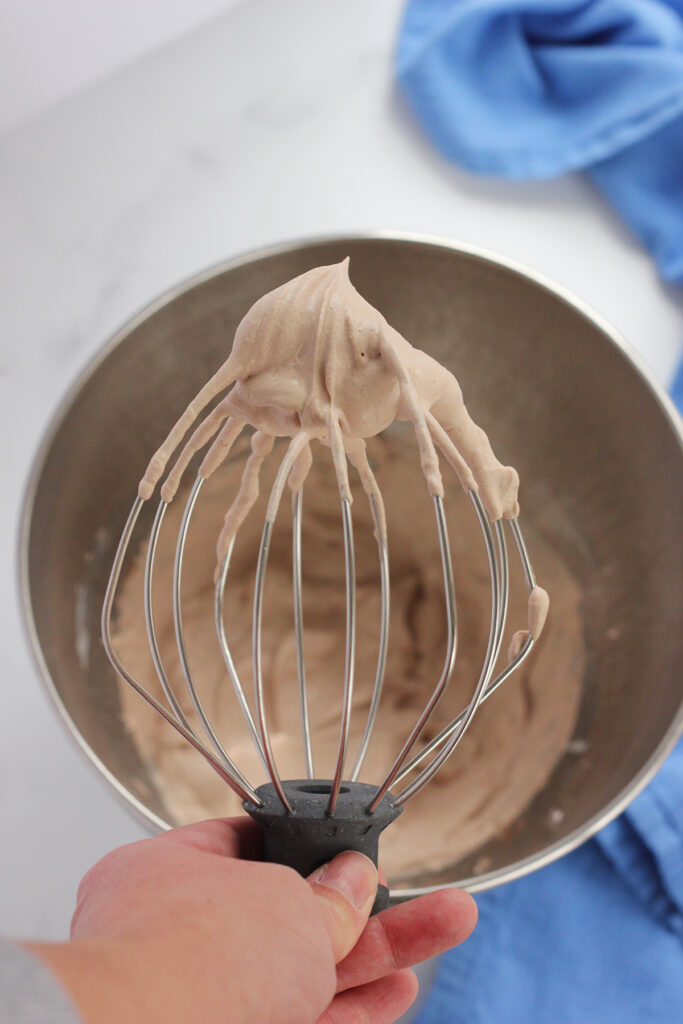 top down image showing the inside of a metal mixing bowl with a whipped chocolate topping with a metal whisk attachment being held over the top