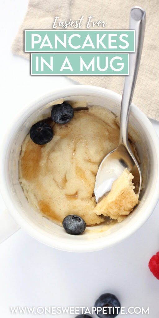 top down image showing a white mug filled with a pancake that is topped with syrup and blueberries. A spoon holds a bite of the pancake. Off to the side are more berries and a tan napkin