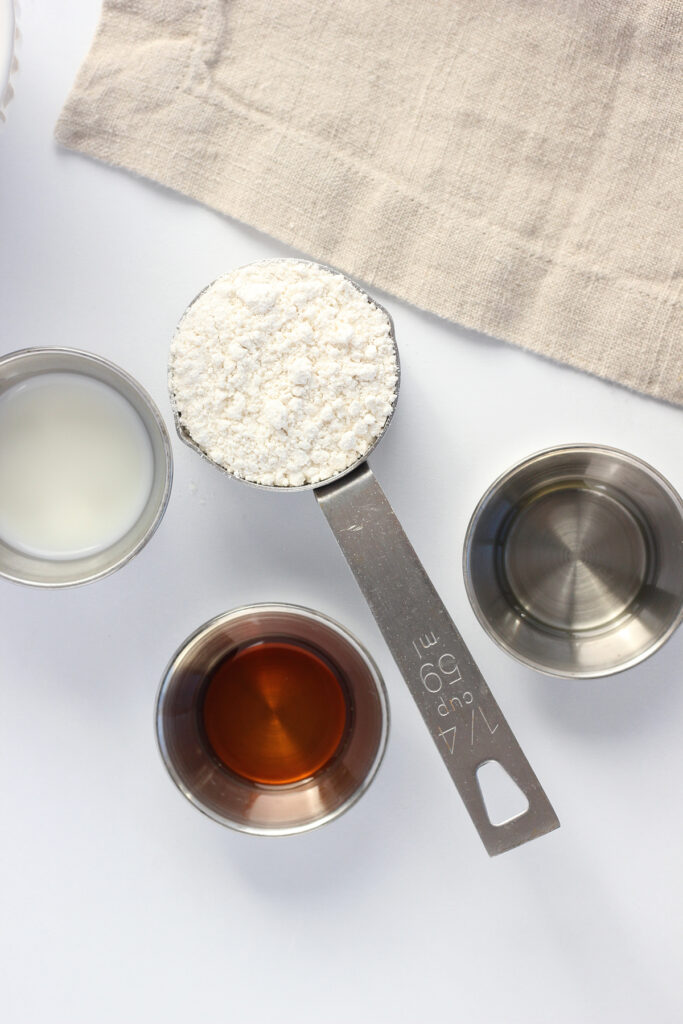 top down image showing a white table top with a 1/4 cup measuring cup filled with pancake mix, and three small metal dishes filled with milk, oil, and vanilla extract. A light tan napkin is off to the side