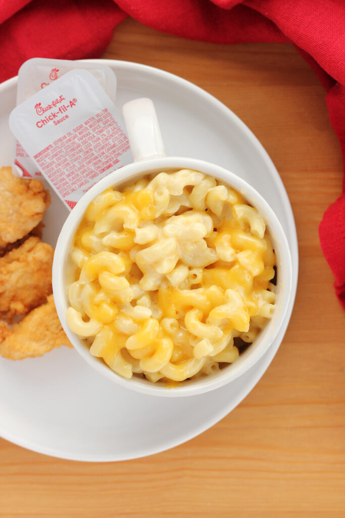 top down image showing a small white mug filled with macaroni and cheese on a white plate with chicken nuggets and two packets of chick fil a sauce