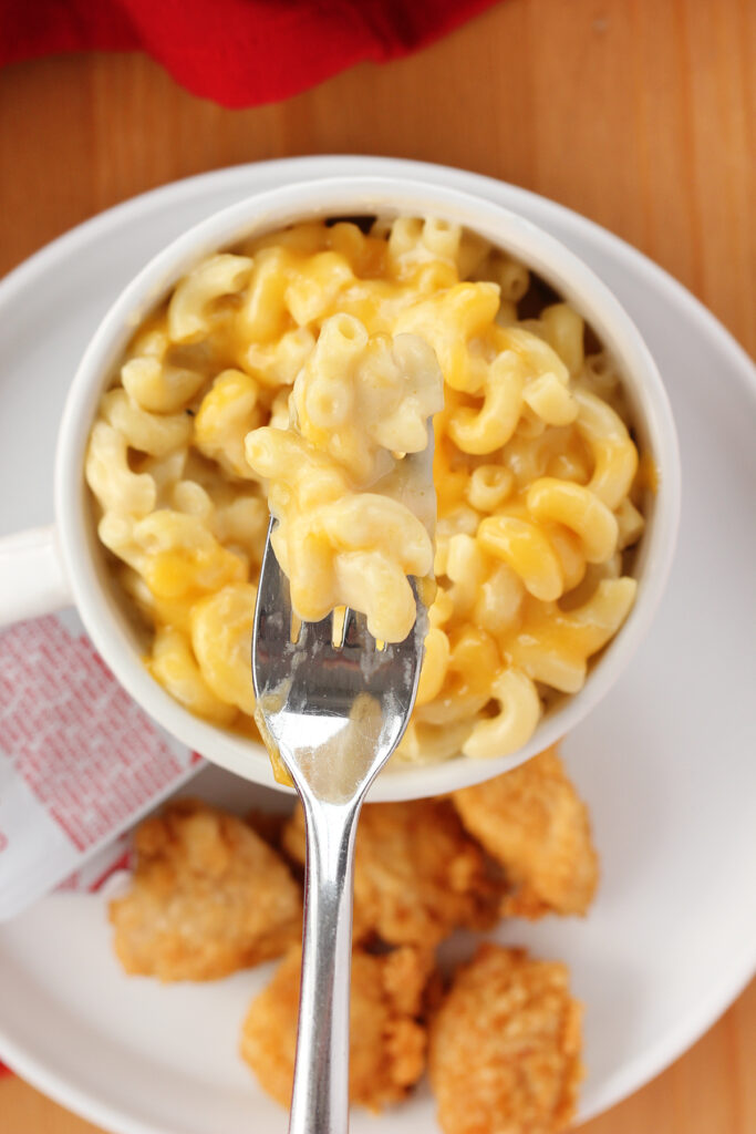 top down image showing a small white mug filled with macaroni and cheese on a white plate with chicken nuggets and two packets of chick fil a sauce with a bite on the fork