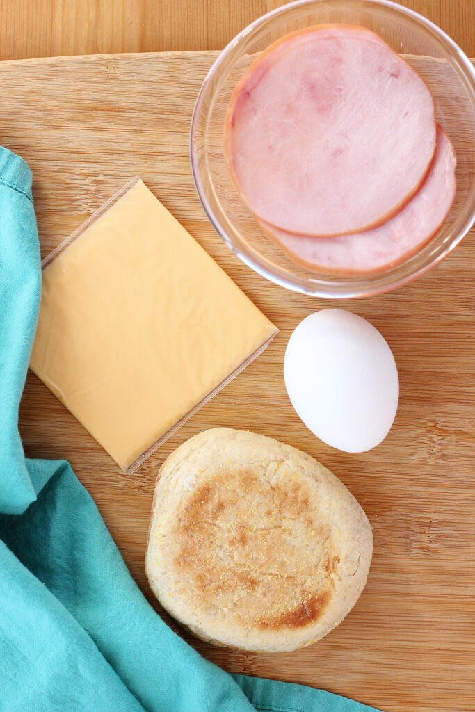 top down image showing an english muffin, egg, slice of american cheese, and two slices of canadian bacon on a wooden table with a teal napkin