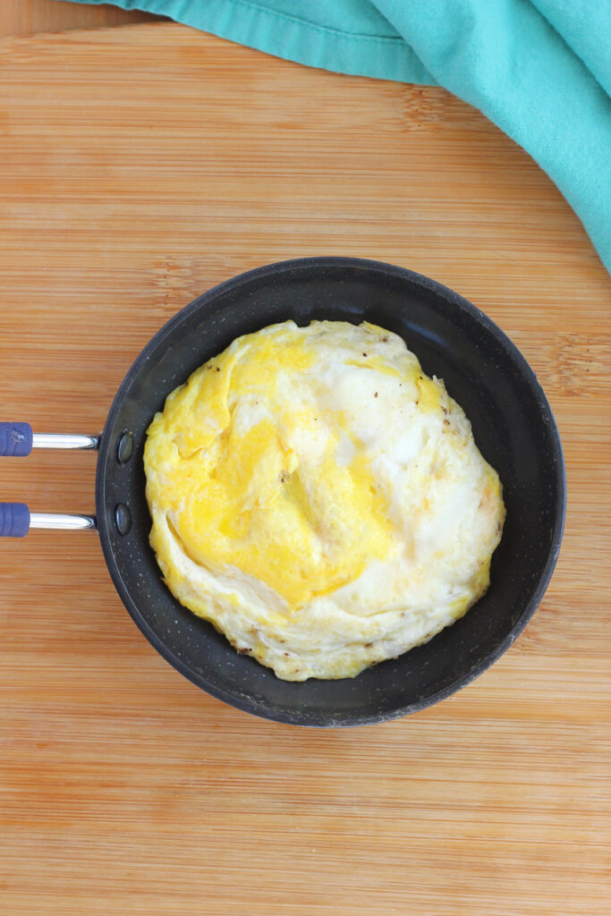 top down image showing a small egg pan with a fried egg inside. The pan is sitting on a wooden counter with a teal napkin off to the side