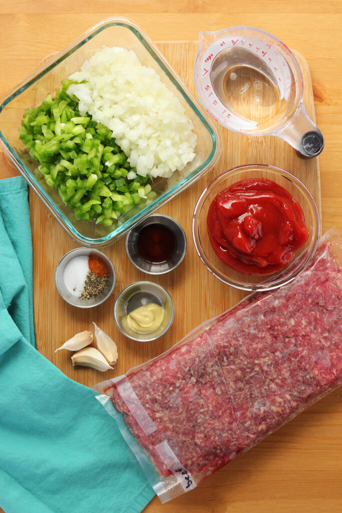 Top down image showing a spread of ingredients on a wooden table including chopped green bell pepper, chopped onion, a measuring cup of water, a bowl of ketchup, three small metal dishes holding a dark brown liquid, seasonings, and mustard, three garlic cloves and a package of ground beef 