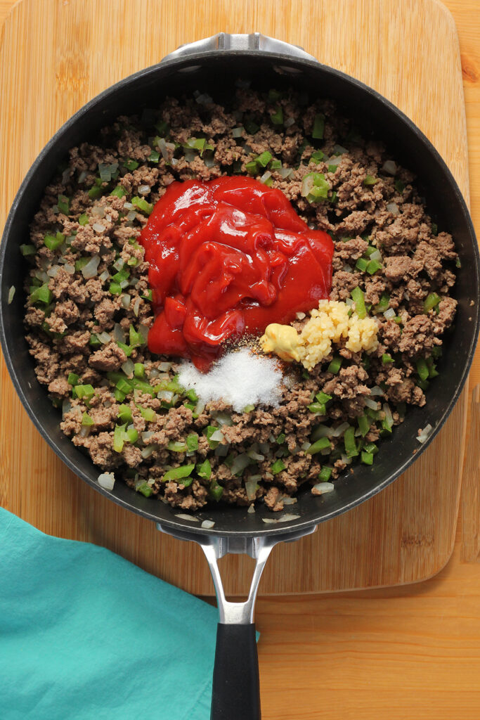 top down image showing a black skillet filled with cooked ground beef and onions topped with ketchup, mustard, garlic and seasonings
