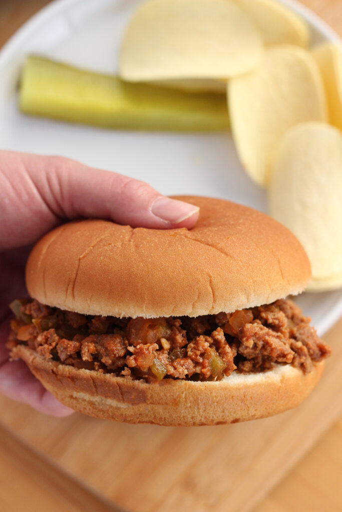 hand holding a bun filled with a ground beef sauce mix hovering over a white round plate that has chips and apickle